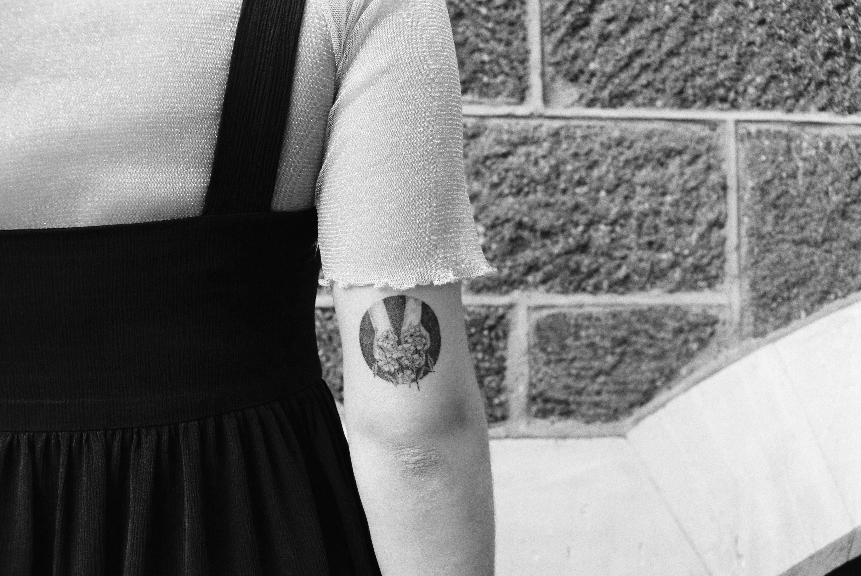 Black and white photo of Bleu's arm and torso showing a circular tattoo of hands holding flowers